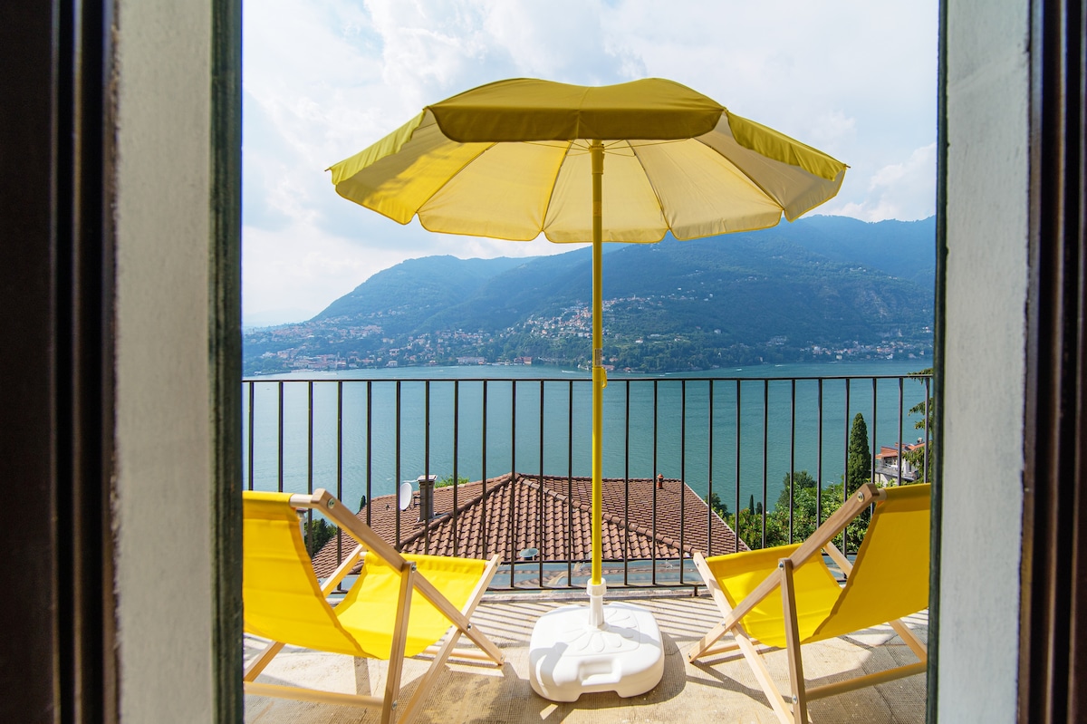 A sunny terrace features a yellow umbrella providing shade over two bright lounge chairs. The view encompasses expansive waters and surrounding mountains, with a glimpse of terracotta rooftops in the foreground, creating a serene outdoor space for relaxation.