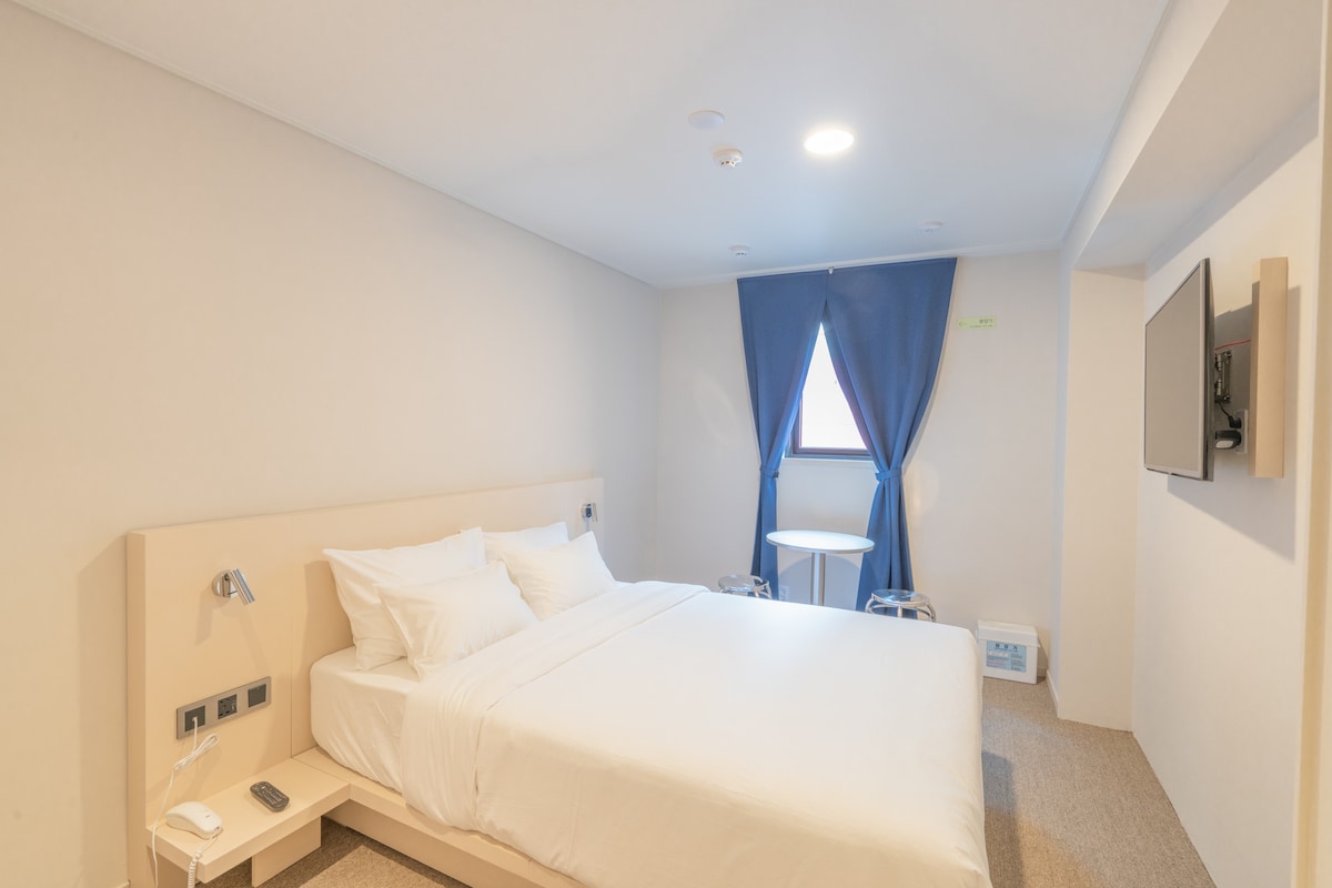 A serene sleeping area features a spacious bed with crisp white linens. A small table is positioned near a window, which is dressed with dark blue curtains. A wall-mounted television and control panel can be seen, contributing to a comfortable atmosphere.