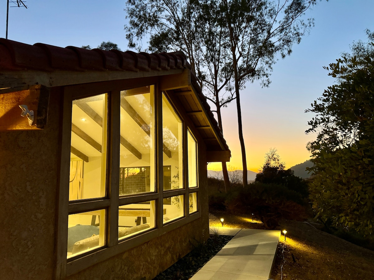 The exterior of the home is framed by tall trees and illuminated with pathway lights. Large windows showcase the inviting interior. A soft sunset glow enhances the natural colors of the surroundings, creating a serene atmosphere.