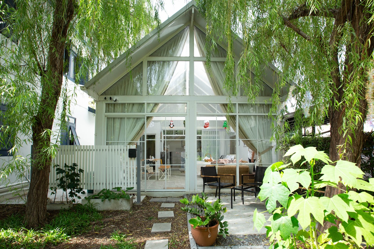 The exterior of the cabin features a high, slanted roof with a large glass front, framed by greenery. A spacious patio area with seating is visible, surrounded by plants and trees, creating a serene setting for relaxation.