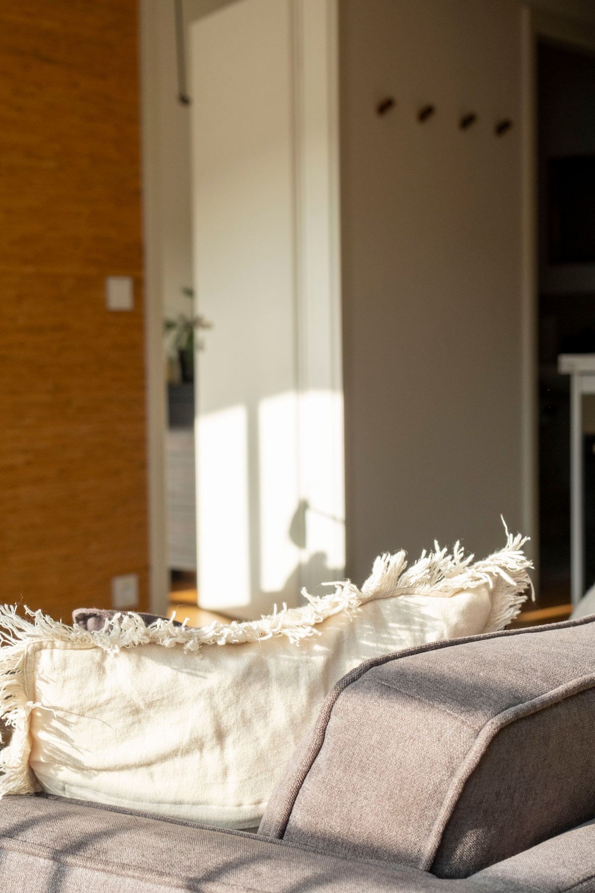 A close-up of a cozy sofa is shown, featuring a textured cushion with fringed edges. Soft sunlight filters through nearby windows, casting gentle shadows against the warm-toned wall and leading towards an open doorway, creating a sense of inviting warmth and comfort.