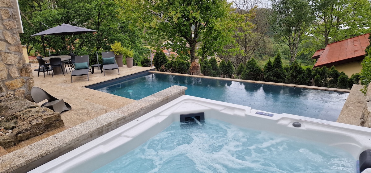 A jacuzzi is situated beside a heated pool, surrounded by a stone patio. Outdoor seating is arranged under a shaded gazebo, with lush greenery and well-maintained landscaping in the background, creating a serene outdoor environment.