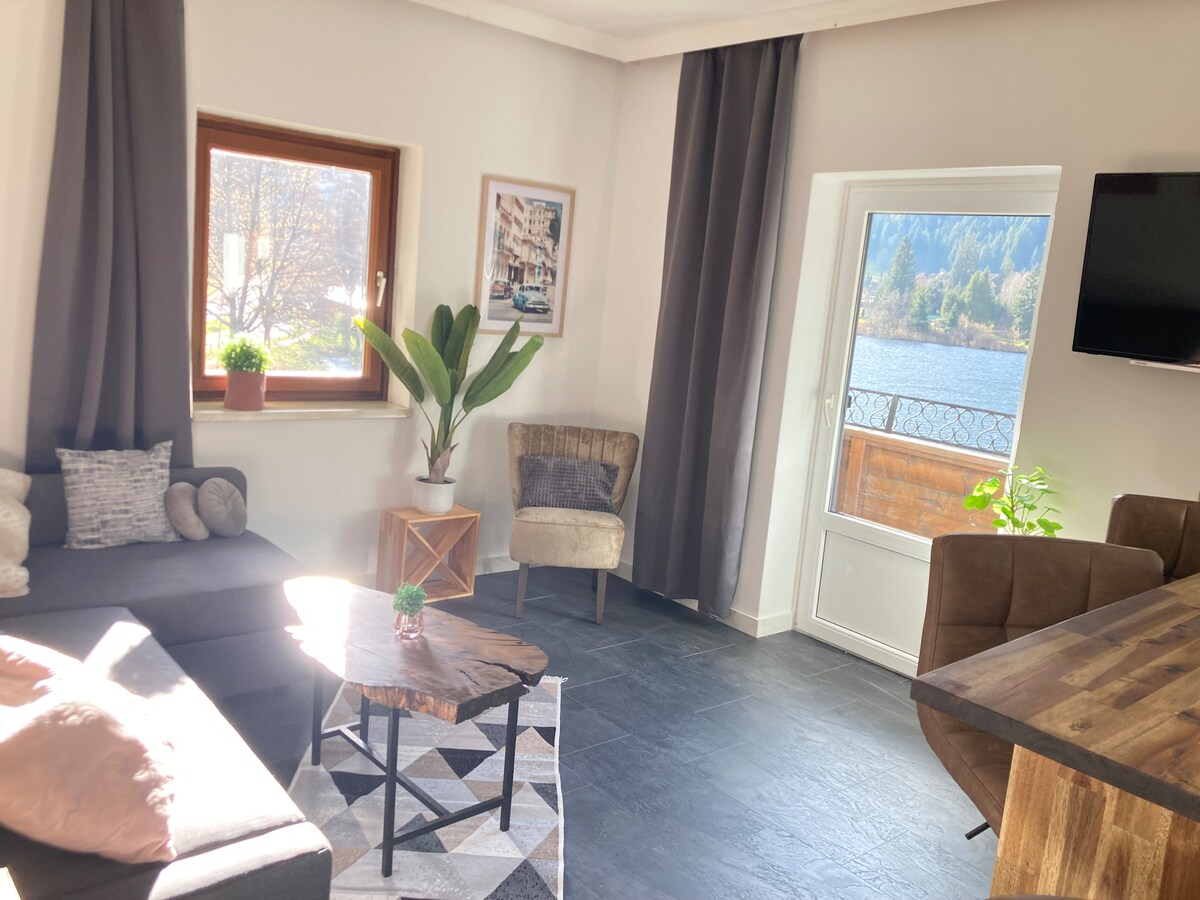 A spacious living area is presented, featuring a large sectional sofa, a round coffee table on a geometric rug, and two accent chairs. Natural light flows in through the large windows, with a view of the lake and mountains visible. Indoor plants add a touch of greenery.