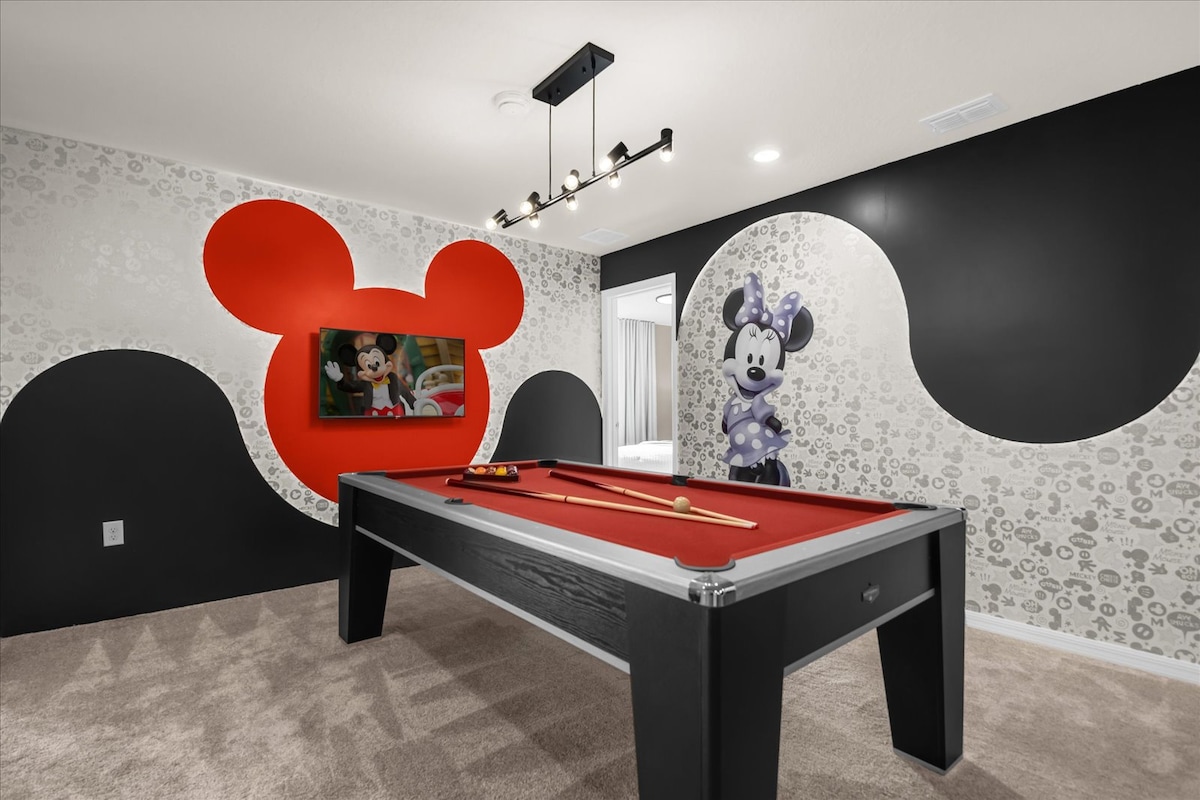 A fun game room features a billiards table at its center, complemented by whimsical wall art showcasing iconic Disney characters. The space is accented with a large red Mickey Mouse silhouette and coordinating designs, set against a contrasting black and patterned backdrop.