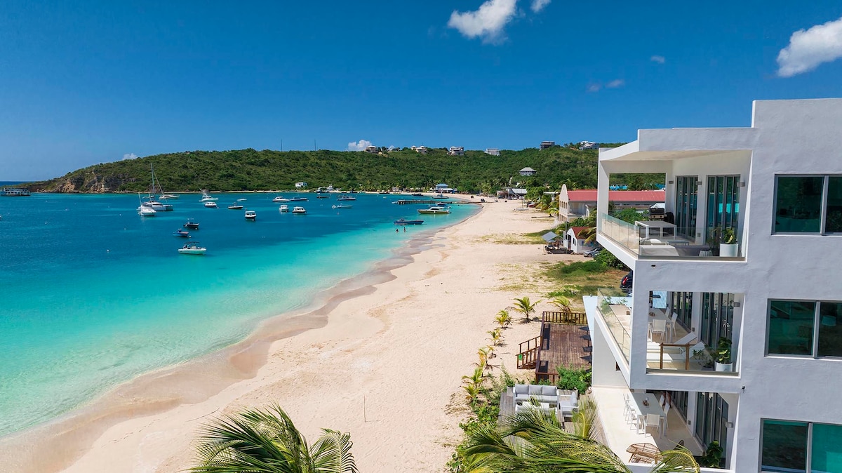 A panoramic view showcases the soft sandy beach meeting clear turquoise waters. The serene shoreline is dotted with boats floating gently in the bay, while a modern beachfront residence is visible on the right, reflecting sunlight through expansive glass windows.