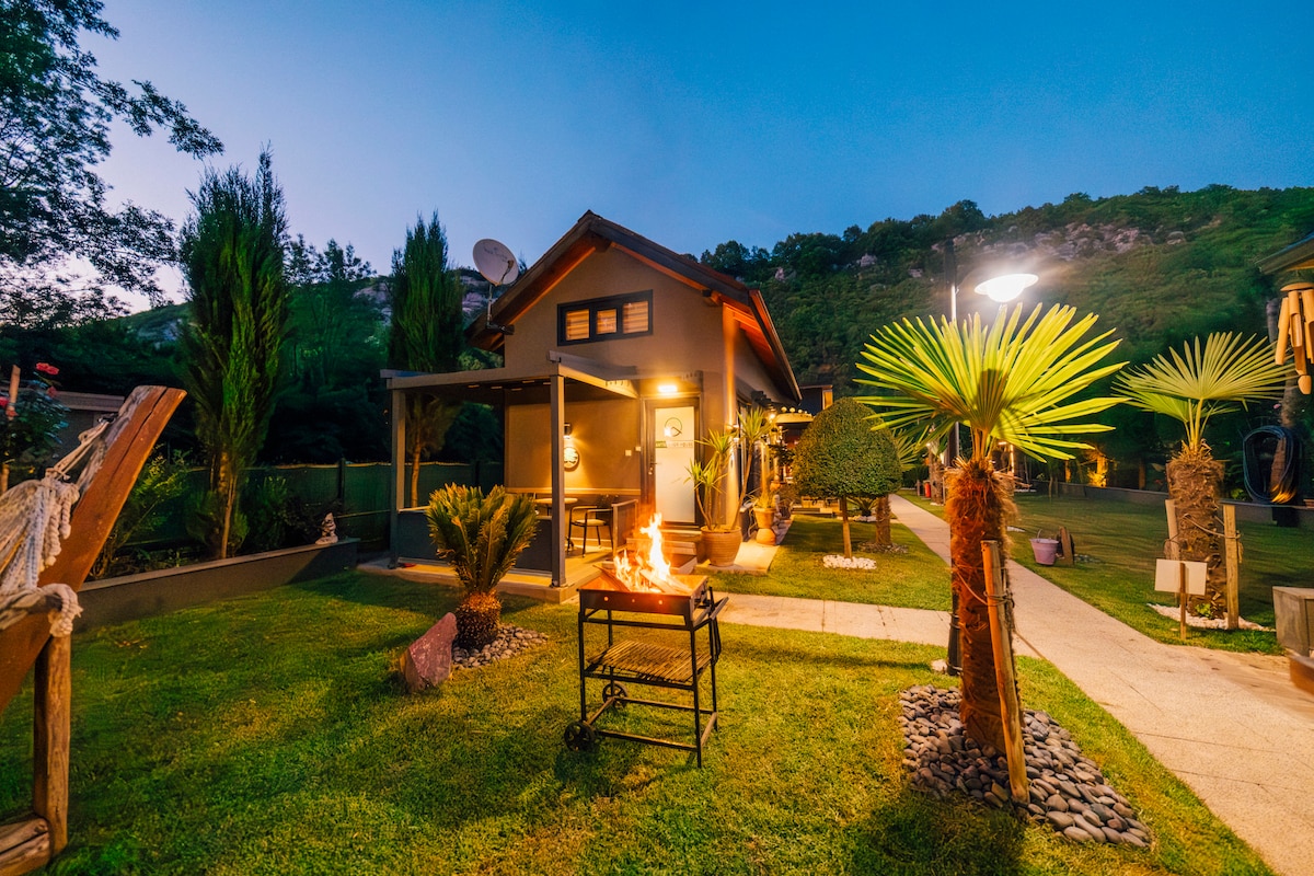 A serene outdoor area features a well-maintained lawn with illuminated pathways, leading to a cozy villa. The space includes seating arrangements and a grill area, surrounded by greenery and decorative plants. Warm lighting enhances the inviting atmosphere of the setting.