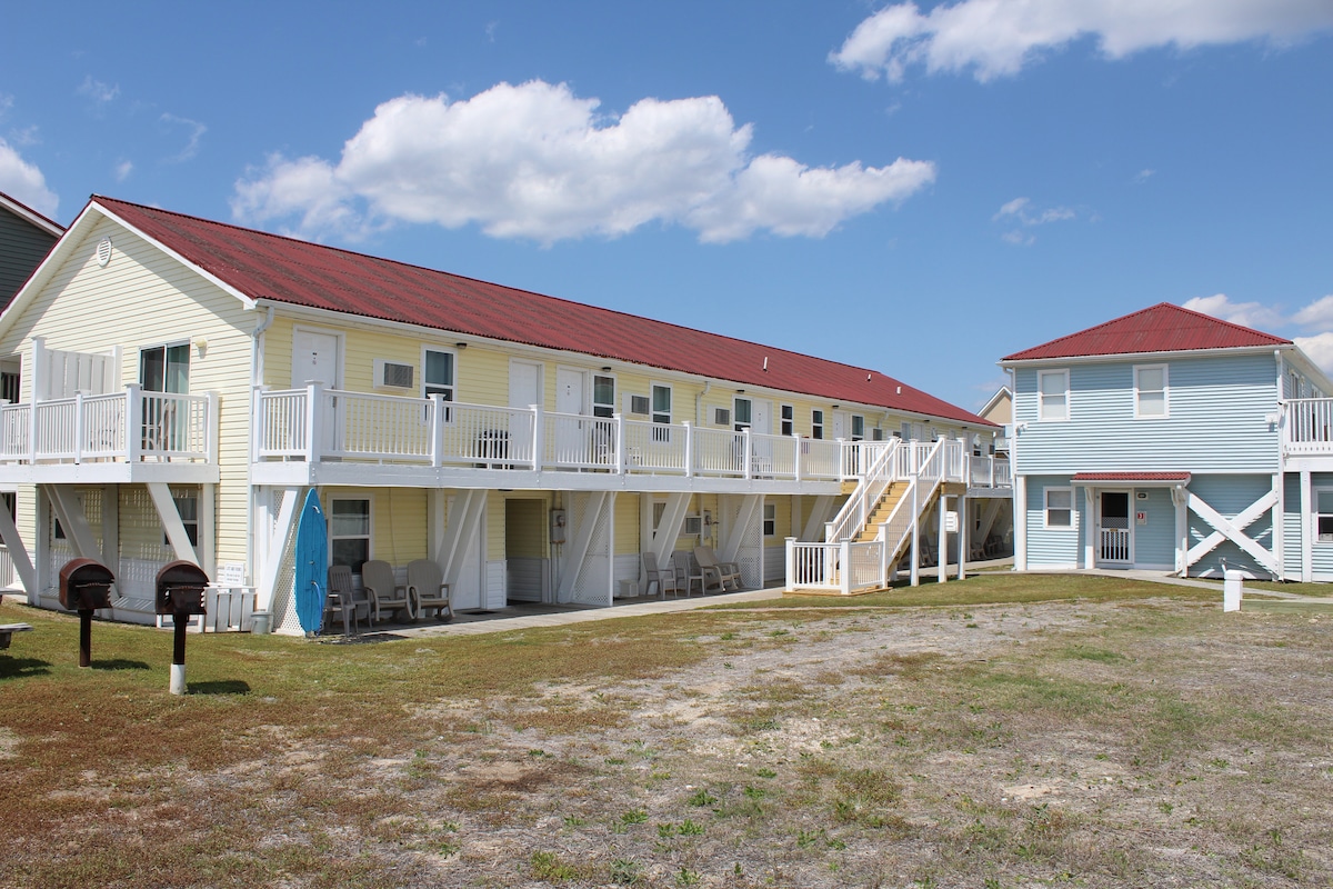 A low-rise building with a pale yellow exterior and a red roof is seen, featuring multiple patio areas with white railings. Friendly outdoor seating is available, along with a pathway leading to a blue building that reflects coastal charm.