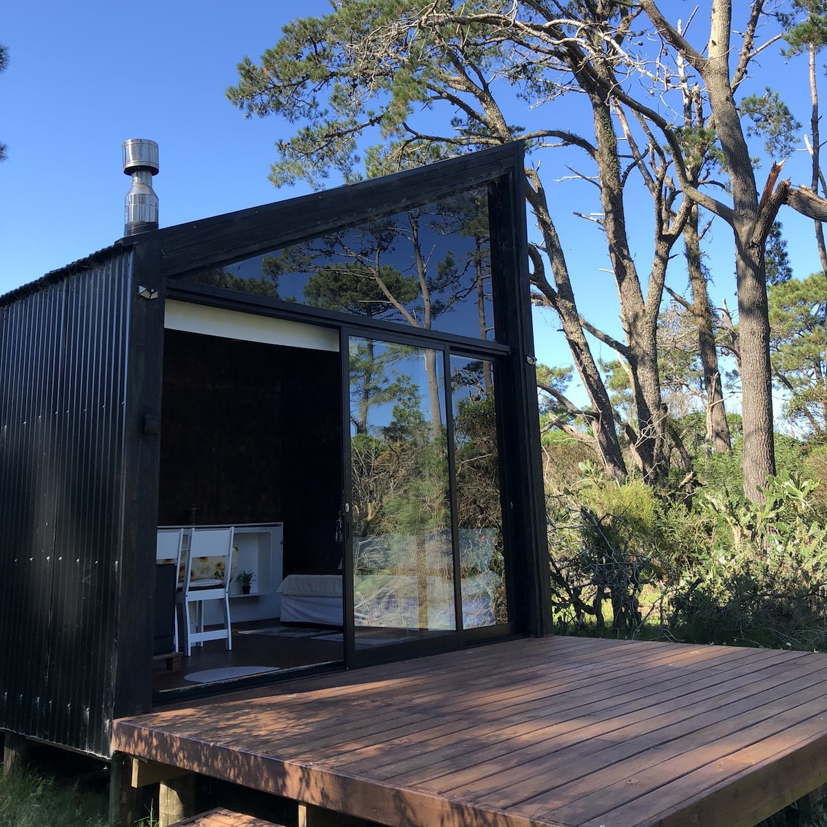A modern cabin is presented with a striking angled roof and large glass doors, opening to a wooden deck. The surrounding trees offer a natural backdrop, enhancing the serene environment. The interior is hinted at through the open space, suggesting a blend of comfort and nature.