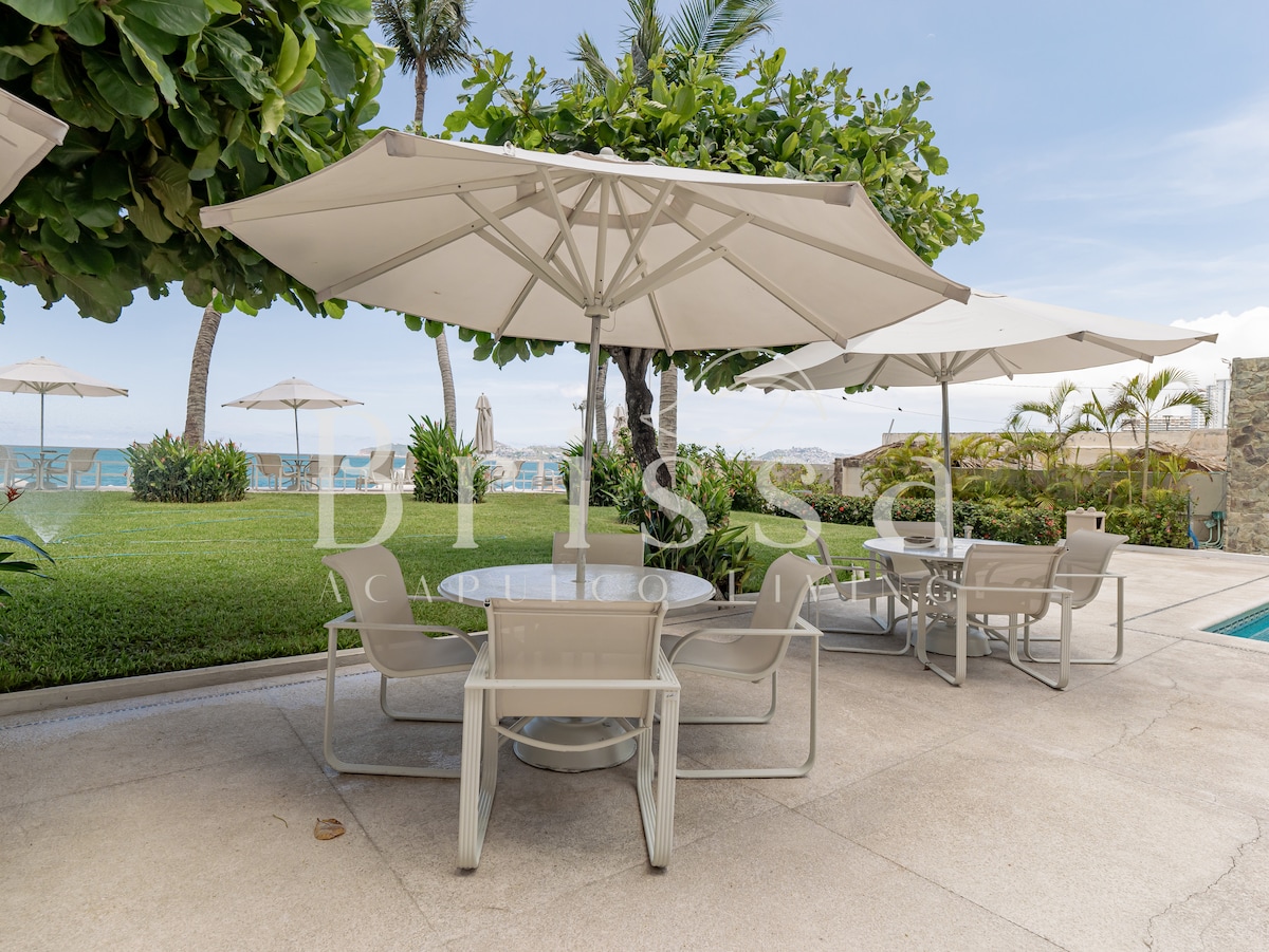 An outdoor area features several light-colored tables and chairs beneath large umbrellas, providing shade. Lush greenery surrounds the space, while views of the ocean can be seen in the background, enhancing the relaxing atmosphere of this coastal setting.