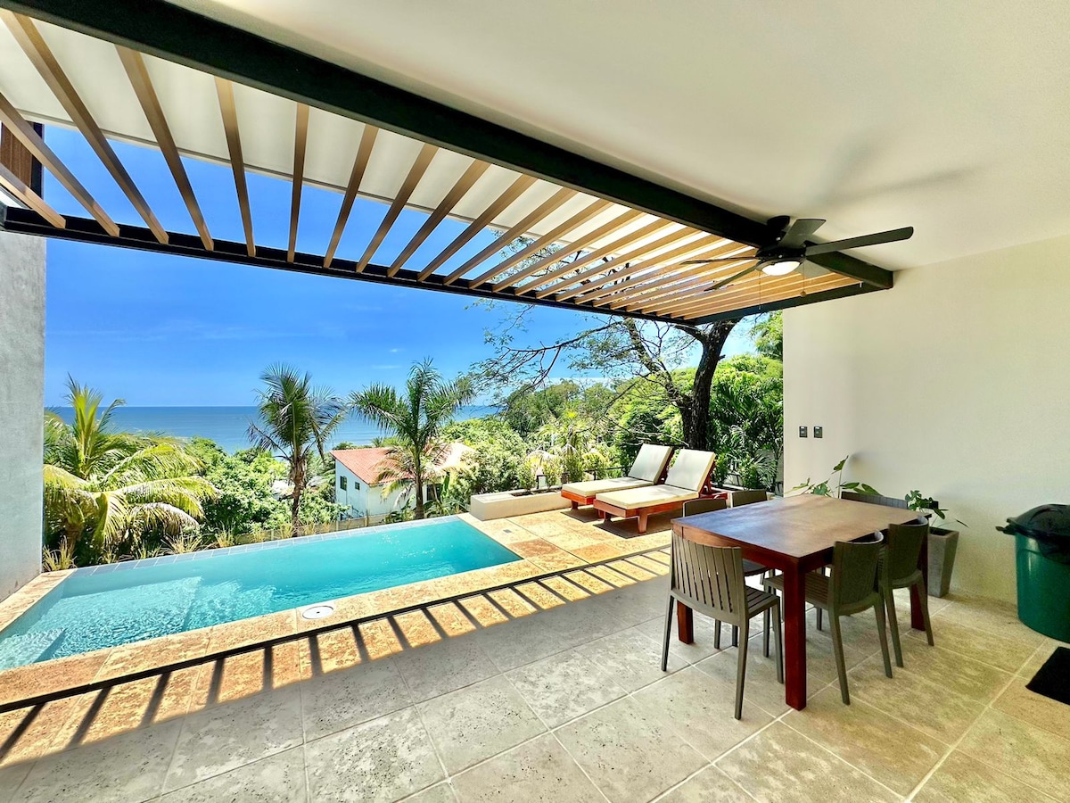 An outdoor lounging area is featured, with a private pool offering ocean views. Sunbeds are positioned beside the pool, while a dining table with chairs is visible. Lush greenery surrounds the space, and large openings provide a connection between indoors and the natural environment.