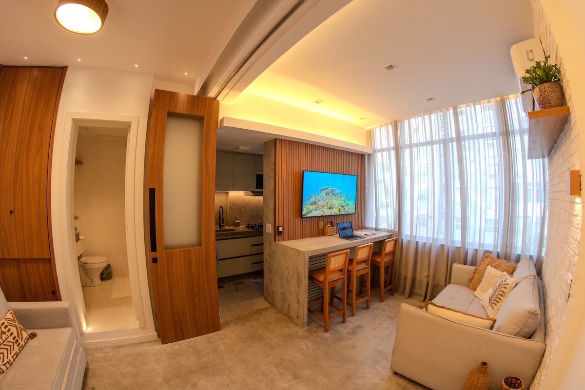 The living area features a comfortable couch and a minimalist wooden table with four chairs, set against a backdrop of large windows covered with sheer curtains. A wall-mounted television displays a vibrant image, while the light wood cabinetry of the kitchen is partially visible.