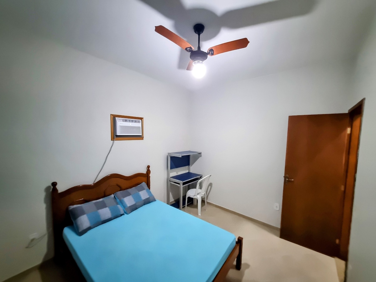 A well-lit bedroom features a spacious bed with blue bedding and two checkered pillows. A small desk and chair are positioned nearby. A ceiling fan is visible, along with a wall-mounted air conditioning unit. A wooden door leads outside.