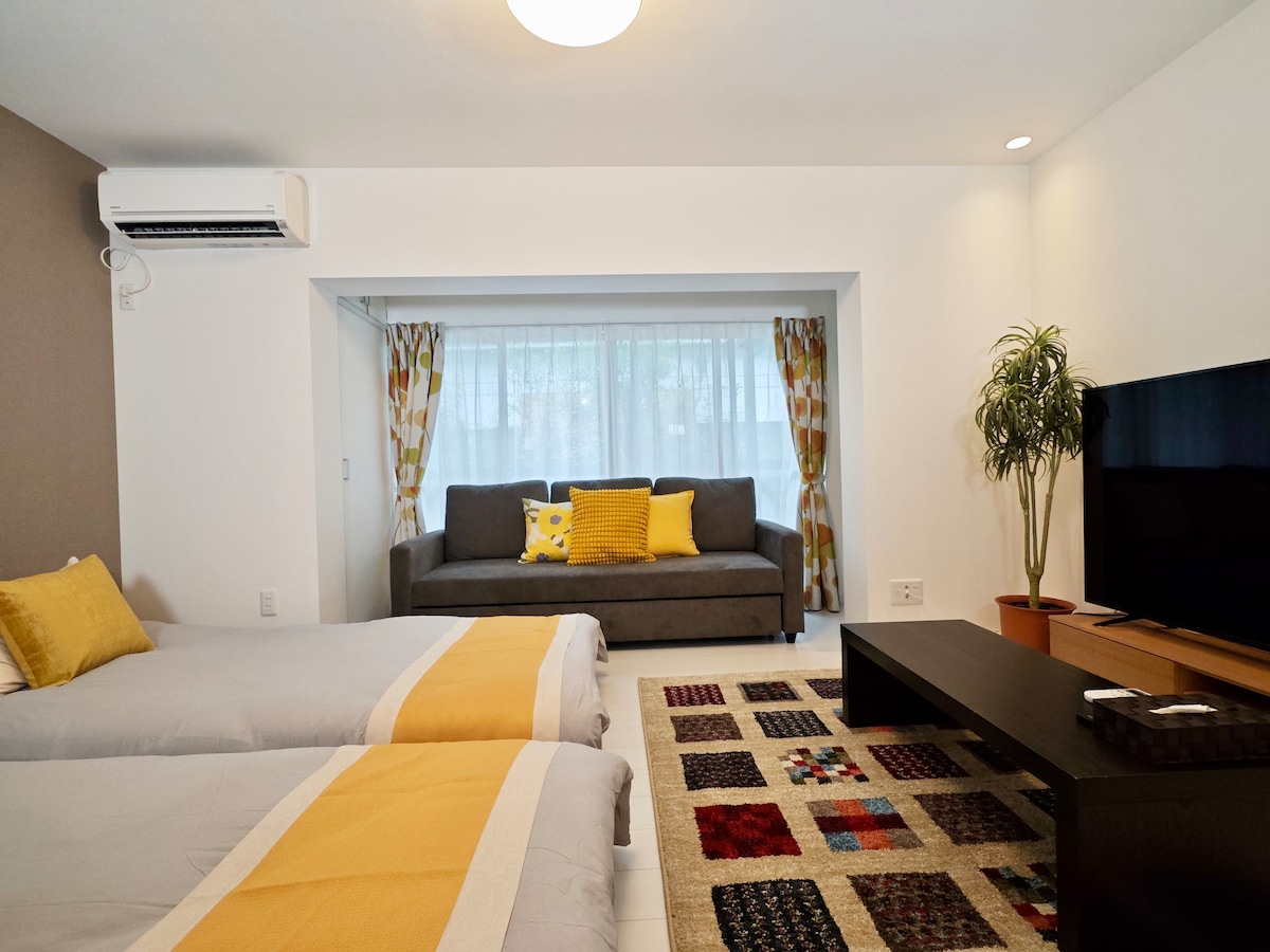 A spacious room is presented with two beds featuring yellow accents and soft linens. A cozy gray sofa adorned with colorful cushions is positioned by a large window with light curtains. A patterned rug adds warmth, while an air-conditioning unit and TV complete the setting.