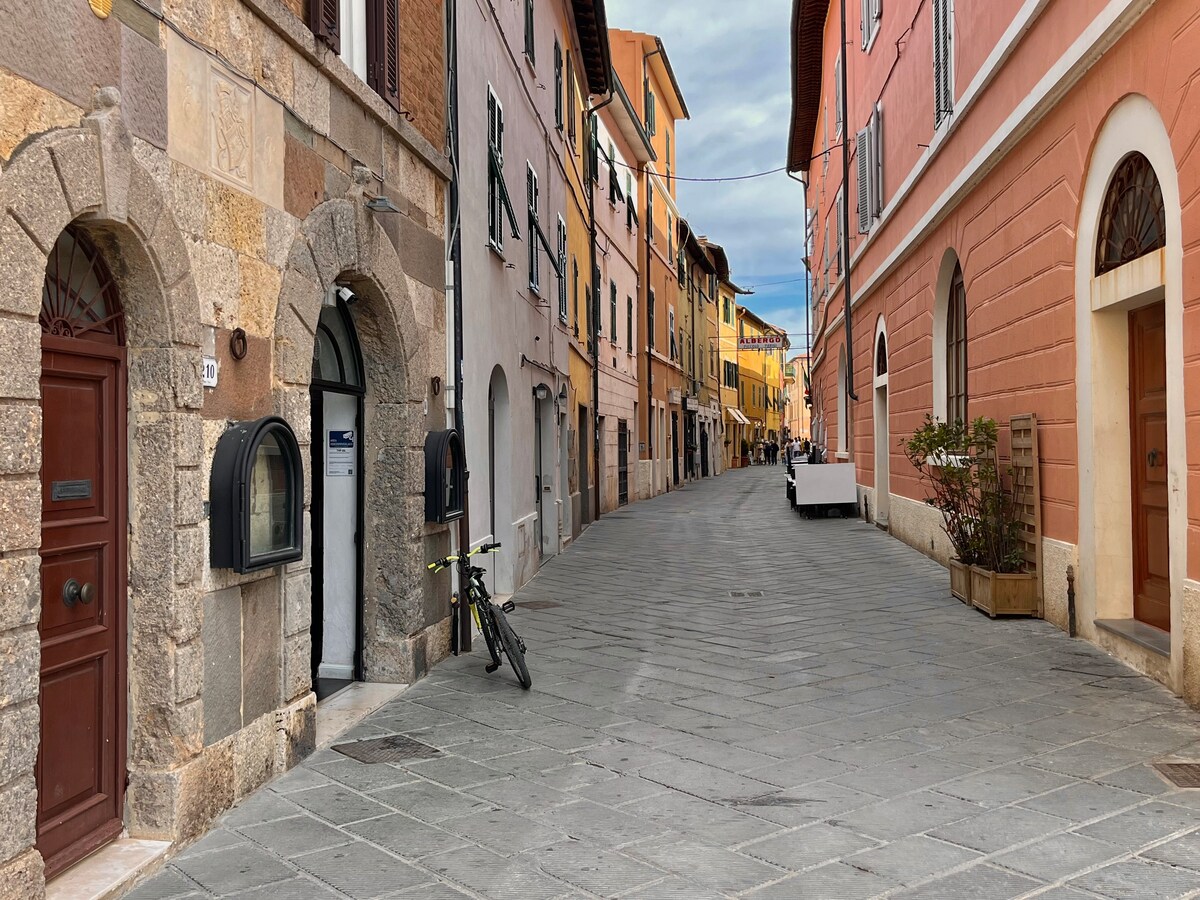 A charming historic street in Orbetello, lined with buildings displaying varied architecture and warm colors. Cobblestone paths lead down the street, with a bicycle parked against the wall. A peaceful atmosphere is observed, accentuated by the absence of heavy traffic.
