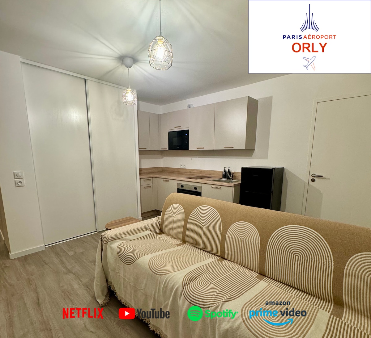 The main living area features a beige convertible sofa that offers a comfortable seating space. A simple kitchenette is visible, equipped with essential appliances and cabinetry. The wall is adorned with modern light fixtures, providing warm illumination.