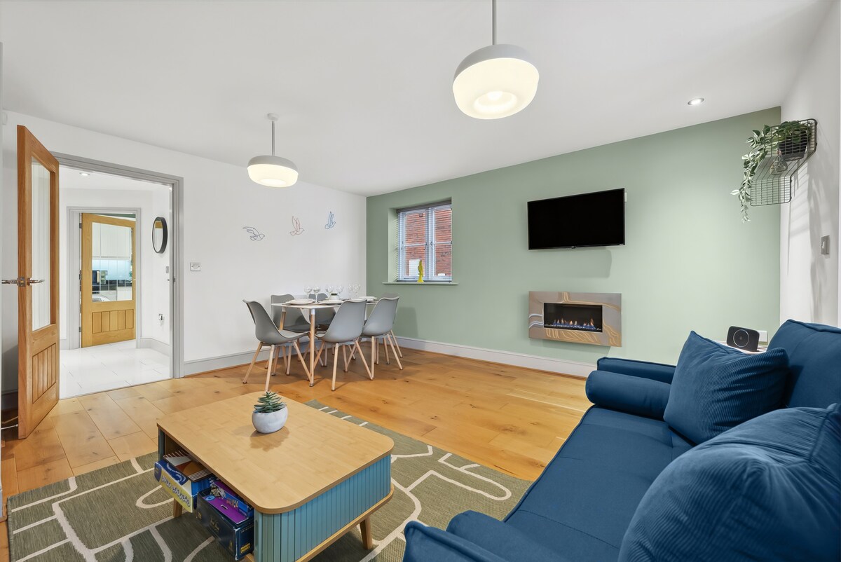 A modern living area is featured with a blue sofa facing a wall-mounted television. A wooden coffee table sits atop a patterned rug, surrounded by a dining area with a table and gray chairs. Natural light enters through a nearby window, illuminating the space.