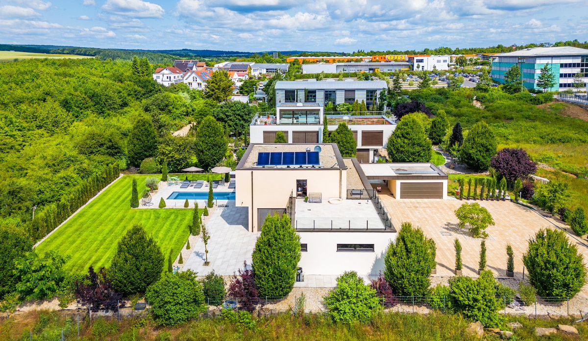 An aerial view of the villa showcases its spacious layout, featuring a well-maintained garden, a swimming pool, and stylish landscaping. The surrounding area is green with trees, while the villa's modern architecture is highlighted against the backdrop of neighboring buildings and the horizon.
