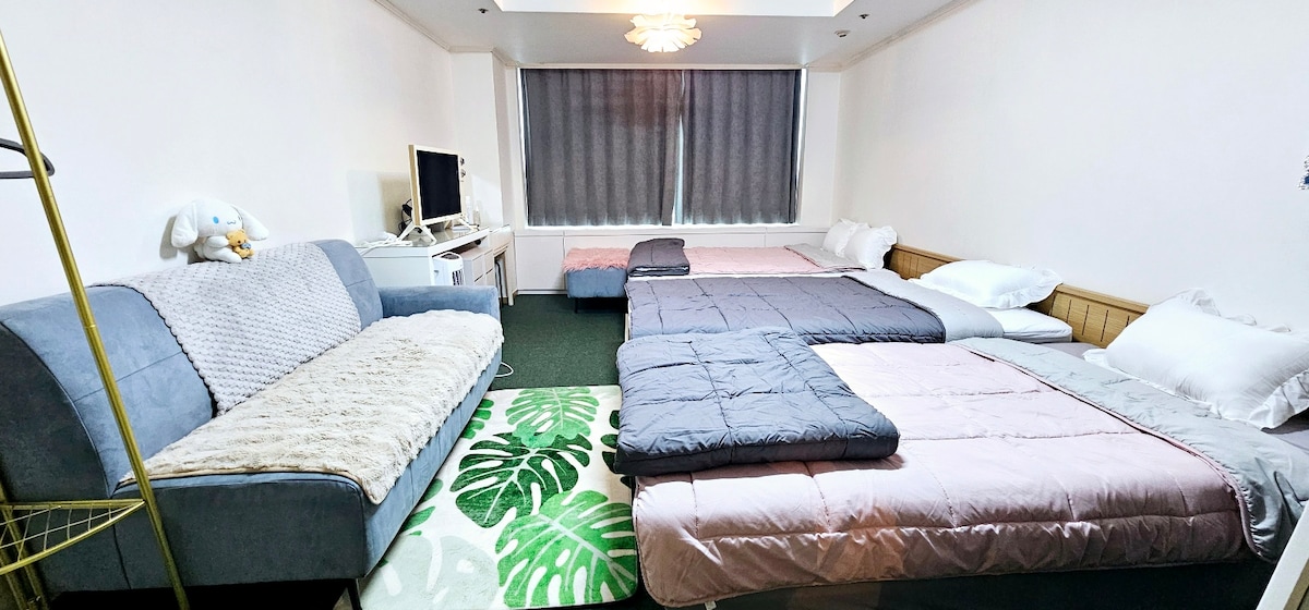 The spacious room features multiple sleeping arrangements, including a queen-size bed and two super single beds. A comfortable sofa sits against the wall, complemented by a decorative rug with green leaf patterns. Light filters through large windows covered with dark curtains.
