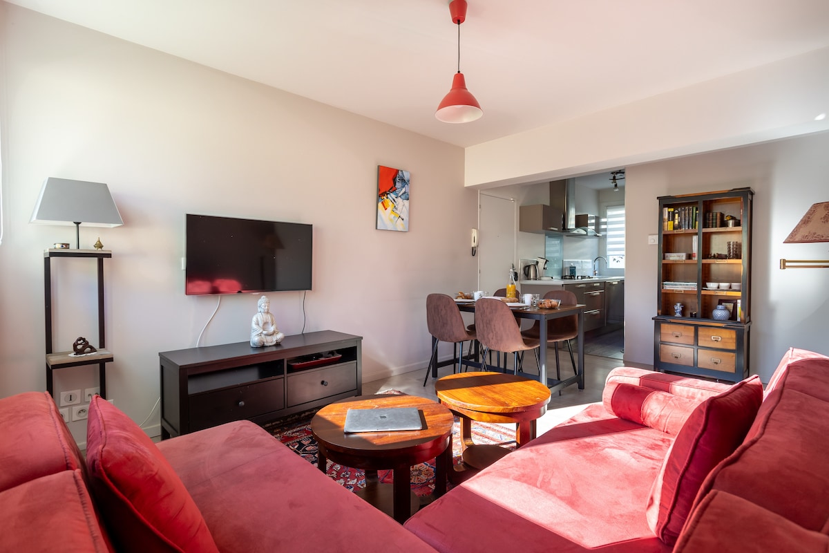 A cozy living area features a large corner sofa in a deep red hue, complemented by two round wooden coffee tables on a patterned rug. A wall-mounted television is visible, and a dining area with four chairs is in the background. Soft, natural light enters through nearby windows.