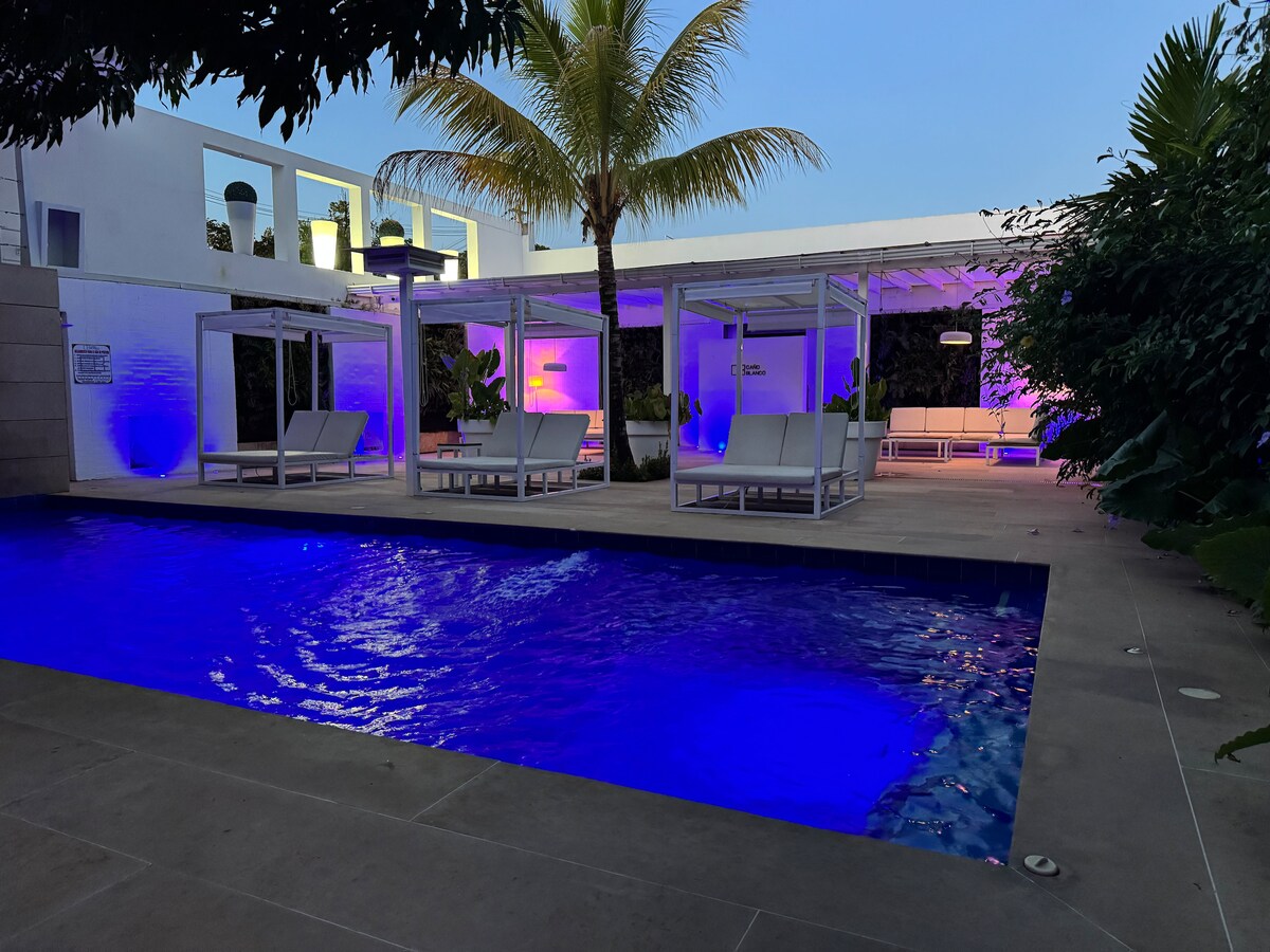 A serene outdoor space features a shimmering pool illuminated in blue. Two lounge areas with white canopies are positioned beside the pool, surrounded by tropical plants. Soft lighting enhances the ambiance as evening approaches, creating a tranquil environment.