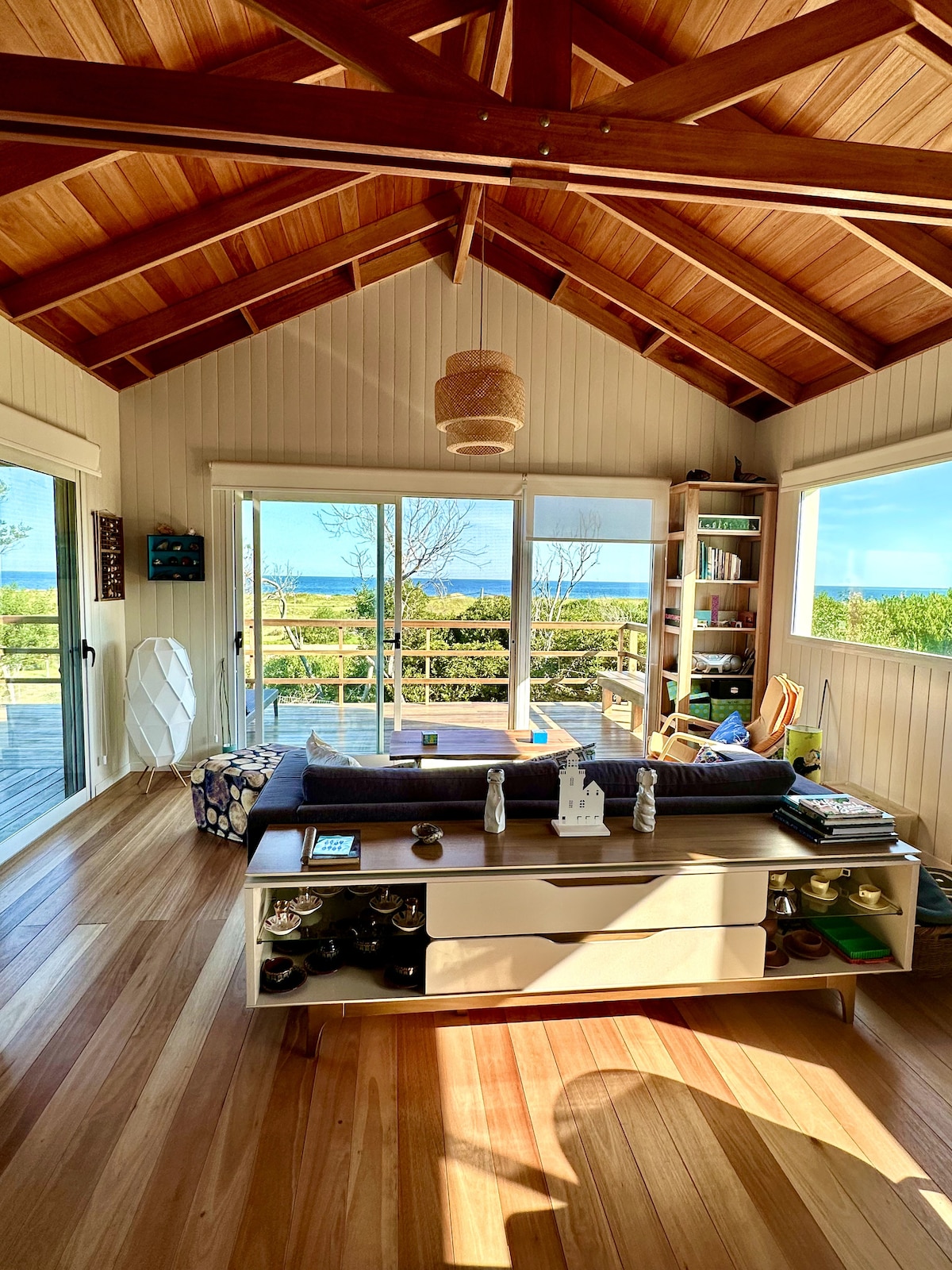 An inviting living area features a high, wooden-beamed ceiling and large glass doors that provide a view of the outdoors. A comfortable seating area is arranged around a low coffee table, while natural light fills the space, highlighting the warm wooden flooring.
