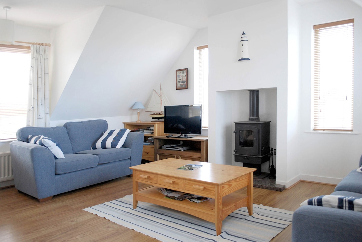 A bright living area is featured, showcasing comfortable blue sofas with striped cushions. A wooden coffee table occupies the center space, positioned atop a striped rug. A television is mounted on a media unit, and natural light is provided by strategically placed windows.