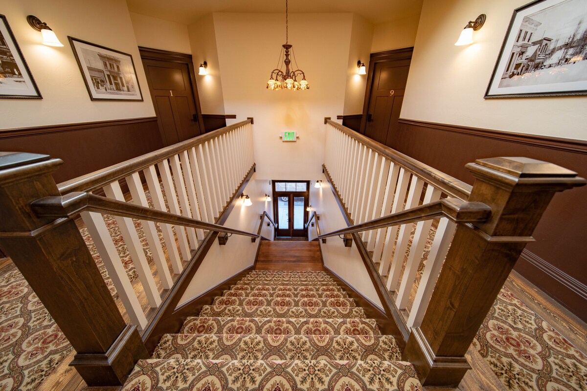 A well-lit staircase features wooden railings and a patterned runner, leading to a welcoming entrance below. Historic photographs adorn the walls, adding character to the space, while a decorative chandelier provides ambient light.