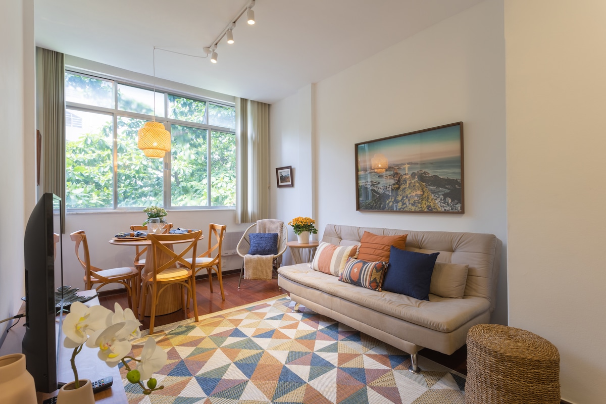 The living and dining area features a light-filled space with a neutral-toned sofa and a round dining table surrounded by four chairs. Decorative pillows add subtle color to the sofa. A large window offers a view of greenery, while a geometric area rug adds texture to the floor.