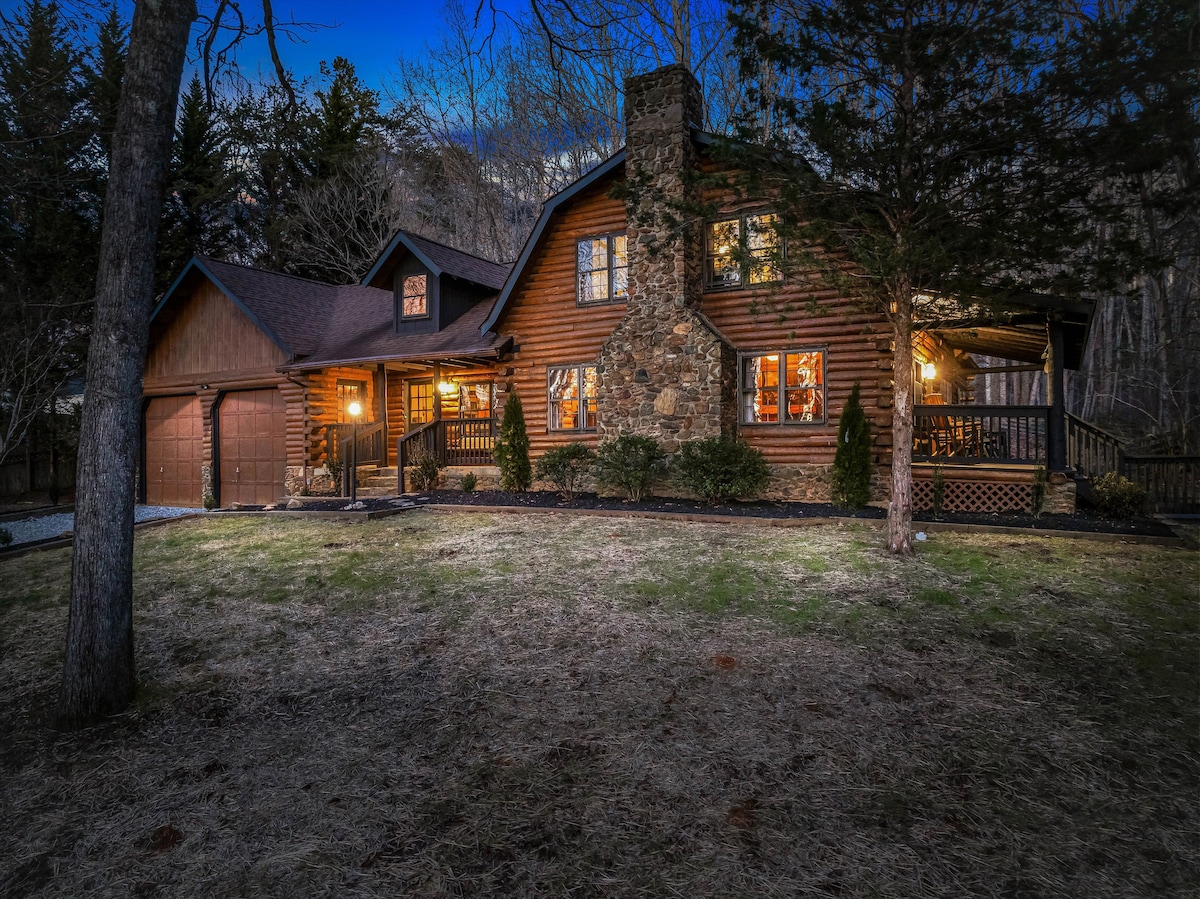 The log cabin exterior is captured at twilight, showcasing the warm glow from the windows. Stone accents on the façade complement the wooden structure, while the surrounding landscape features gently sloping grass and trees that create a serene atmosphere.