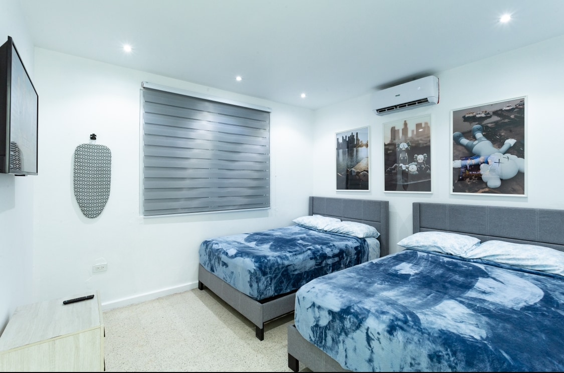 A well-lit bedroom features two spacious beds, each covered with coordinating blue and white bedding. A television is mounted on the wall across from the beds. Three framed artworks add color to the room, and a large window with blinds allows for natural light.