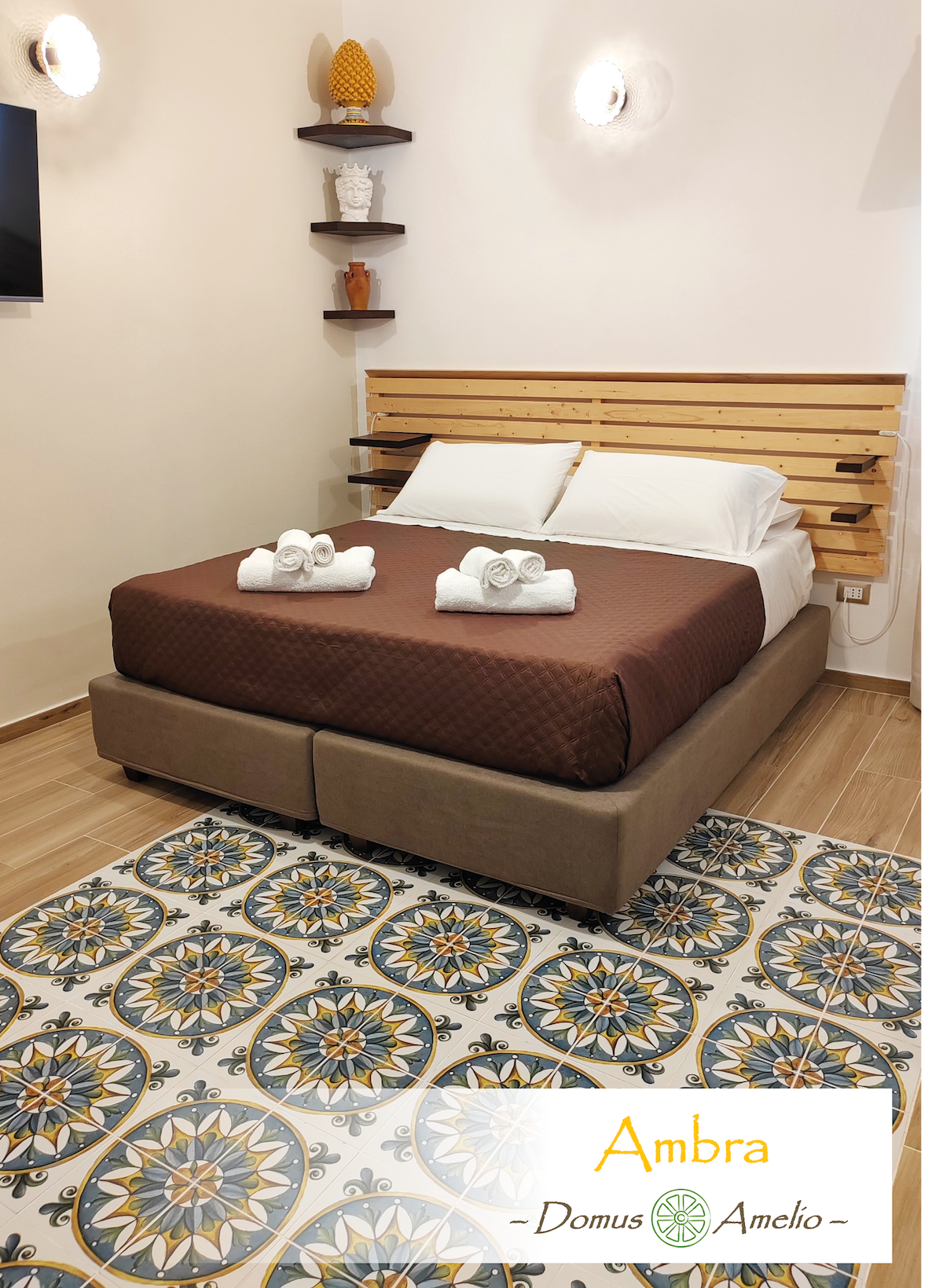 A cozy studio apartment features a spacious bed with a brown quilted coverlet and neatly arranged towels. The room is accented by a colorful tiled floor, and wooden shelves enhance the warm decor. Soft lighting is provided by wall fixtures, creating a calming ambiance.