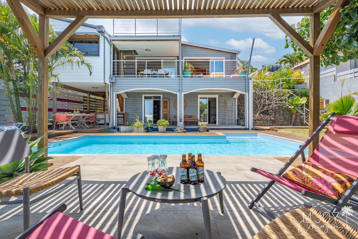 A private outdoor area features a swimming pool, surrounded by tropical plants and shaded by a pergola. Comfortable lounge chairs are positioned near the water. In the foreground, a table is set with two drinks and a small platter, inviting relaxation and enjoyment.
