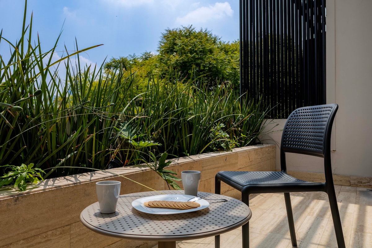 A private balcony is depicted with a small round table and a single chair. Two cups are placed on the table beside a plate with a cookie. Lush greenery is visible in the background, framed by a modern black railing.