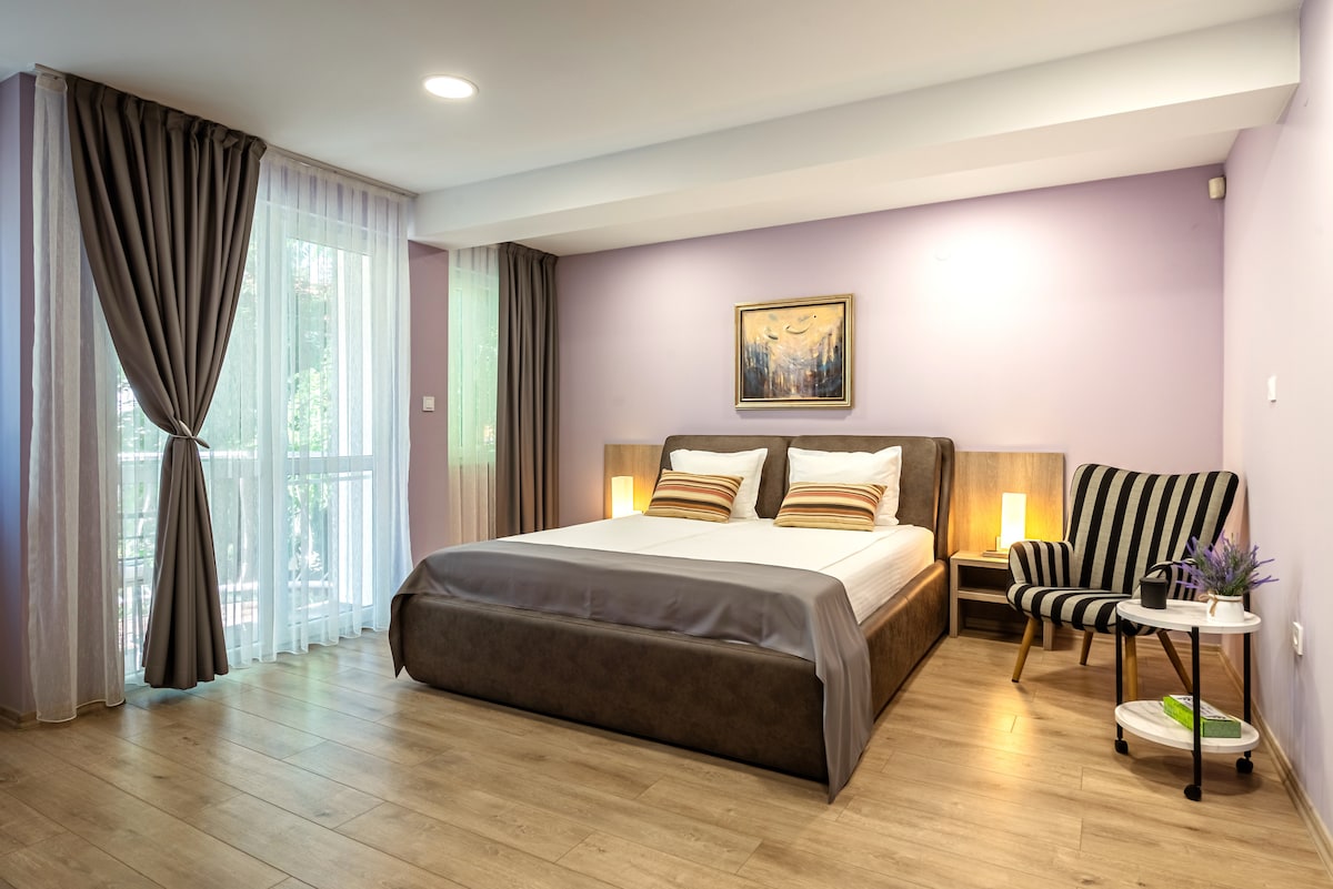 The spacious sleeping area features a comfortable bed with crisp linens, complemented by two bedside lamps. A cozy armchair and side table provide a seating option. Natural light filters through sheer curtains, illuminating the pastel purple walls and enhancing the serene environment.