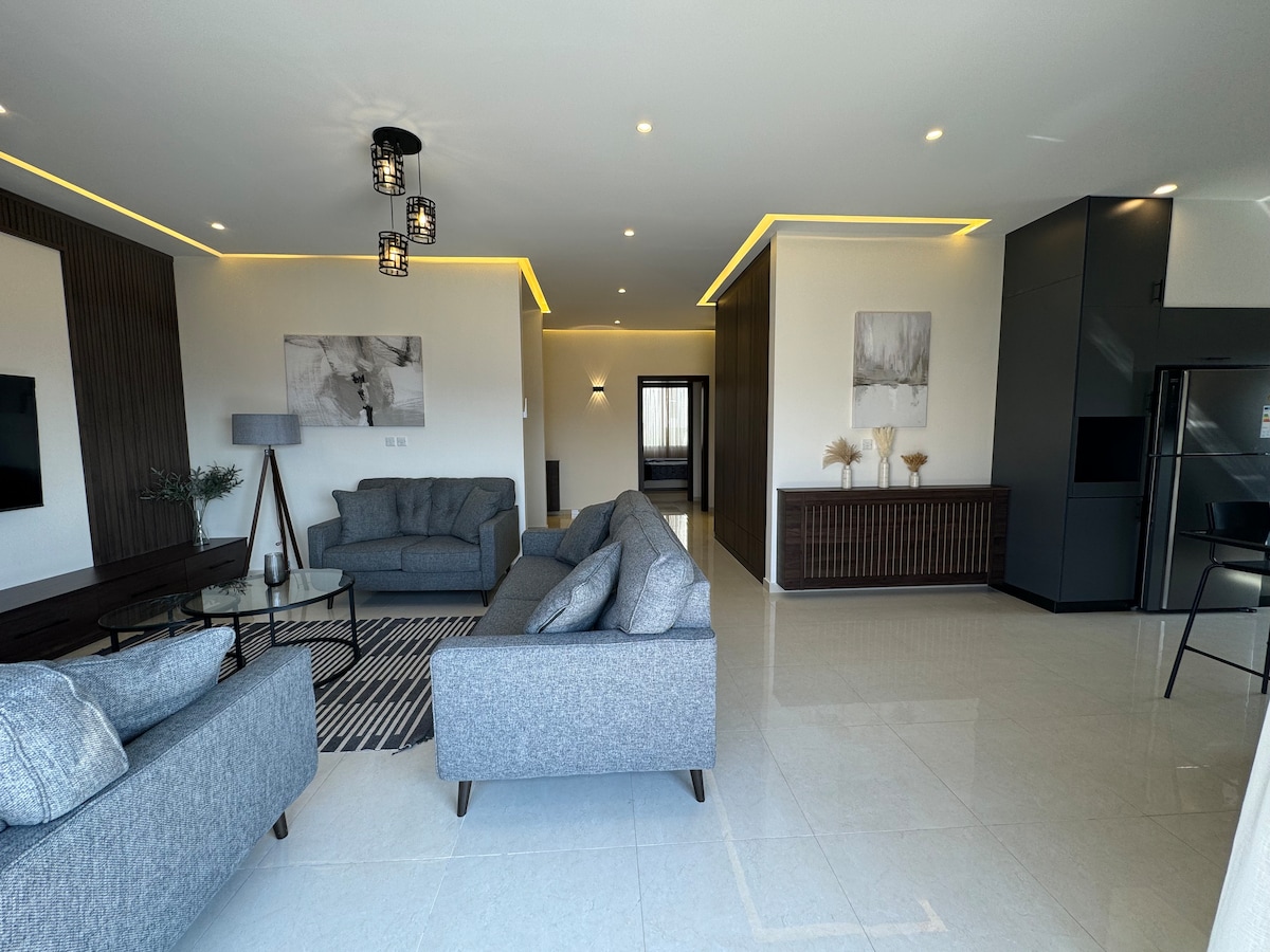 A bright living area features two gray sofas arranged around a geometric rug, alongside a dark wood console. Modern artwork decorates the walls, and a sleek kitchen area is visible in the background, complemented by contemporary lighting.