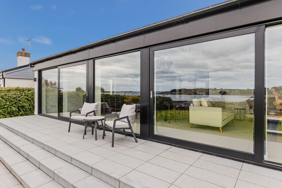 A spacious patio area is shown, featuring two modern outdoor chairs facing large floor-to-ceiling windows. The view displays lush greenery and water beyond, indicating a serene seaside environment. A light-colored sofa can be seen reflected in the glass, enhancing the open atmosphere.