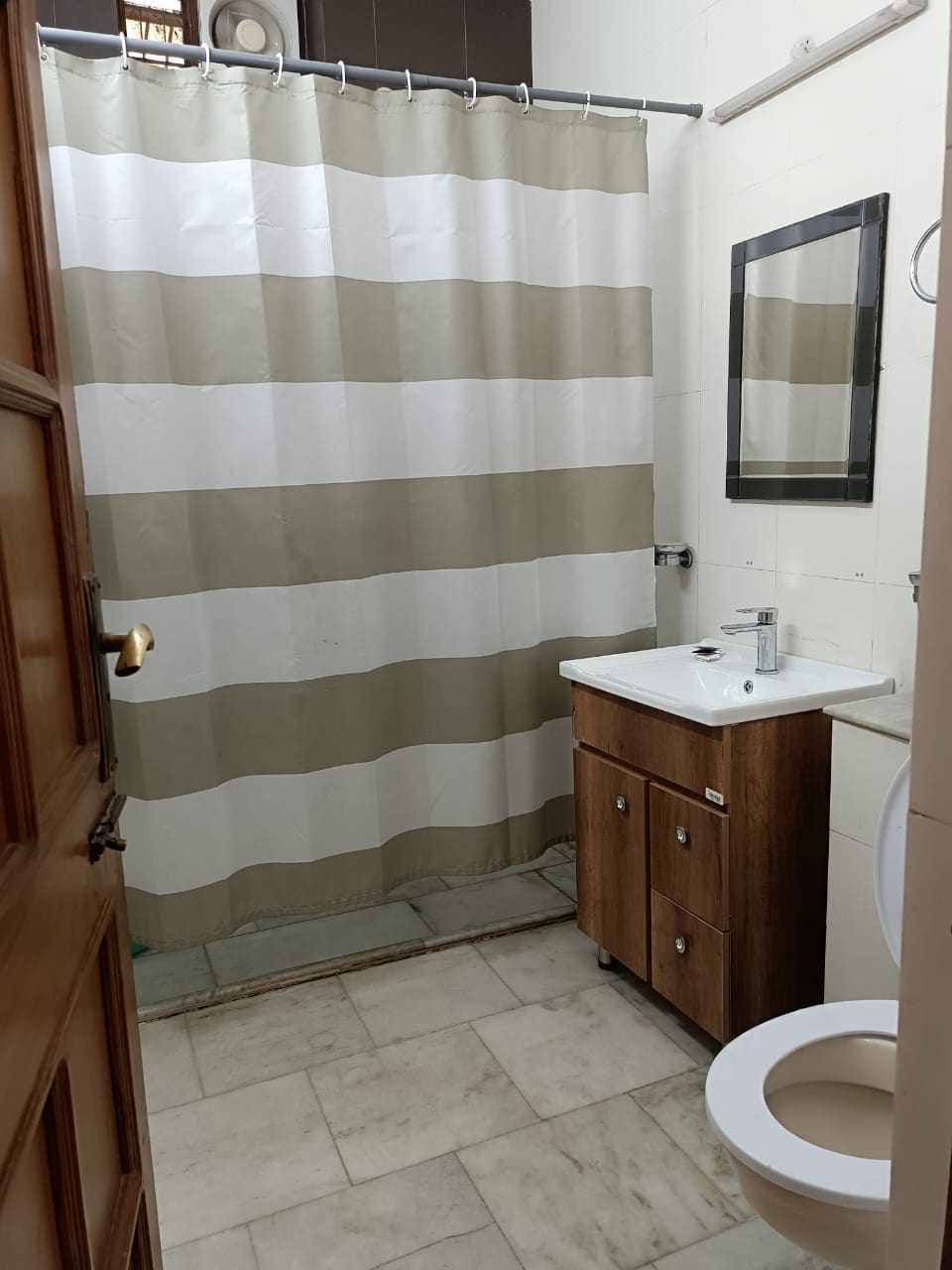 A clean bathroom features a shower with a striped curtain, offering privacy. A wooden vanity with a sink is positioned against a tiled wall, accompanied by a round mirror. Light-colored tiles extend across the floor, contributing to a fresh and open feel.