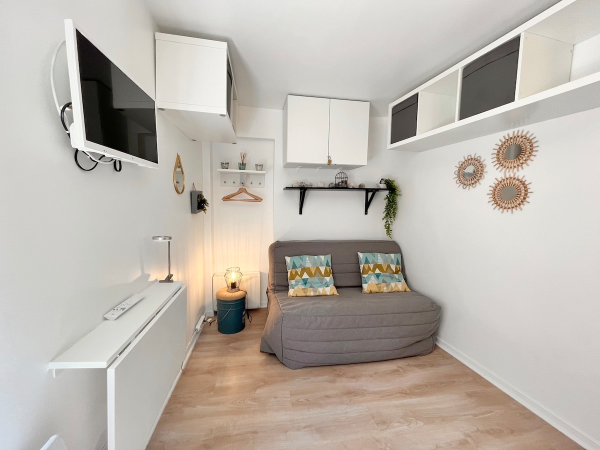 A compact living area is showcased, featuring a gray sofa adorned with patterned cushions. Above, shelves hold decorative items. A wall-mounted television is positioned for easy viewing, while a lamp and small table add practicality to the space.