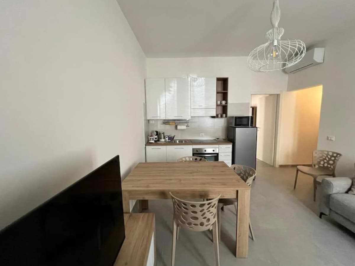 A modern dining area is presented, featuring a light wooden table surrounded by four structured chairs. A compact kitchen is visible in the background, equipped with essential appliances. A television stands on a low unit, and an armchair adds comfort to the inviting space.