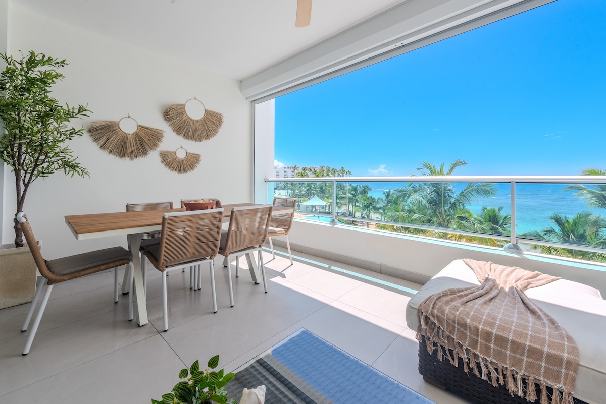 A dining area is set beside a large glass door, offering ocean views. A wooden table is surrounded by several light-colored chairs. Decorative wall hangings add texture to the white walls, while a cozy blanket is draped over a nearby chaise lounge.