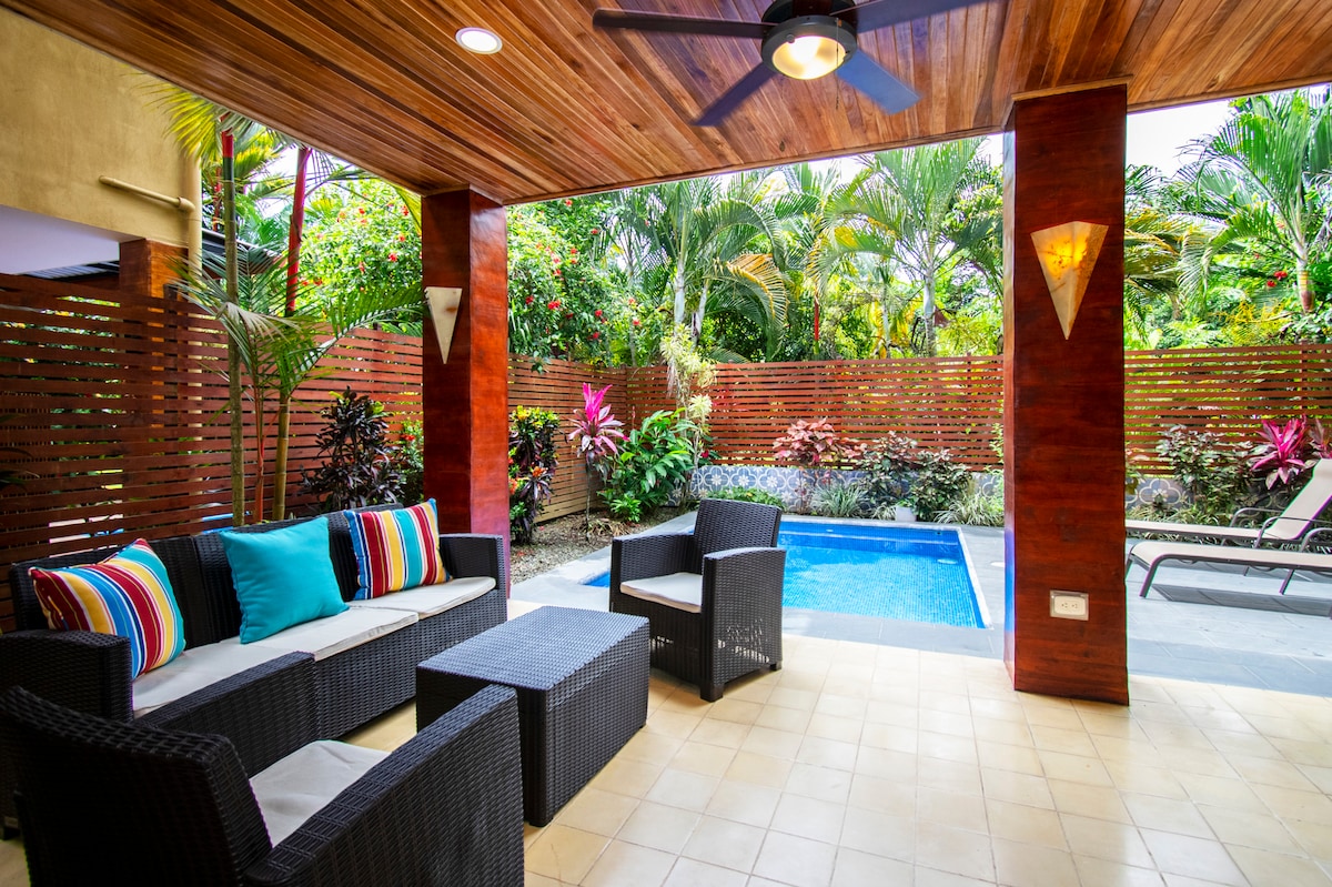 A covered outdoor seating area is highlighted by comfortable furnishings, including a sofa and coffee table. A refreshing pool is visible nearby, surrounded by lush greenery and tropical plants. Soft lighting fixtures are mounted on the wooden walls, enhancing the serene environment.