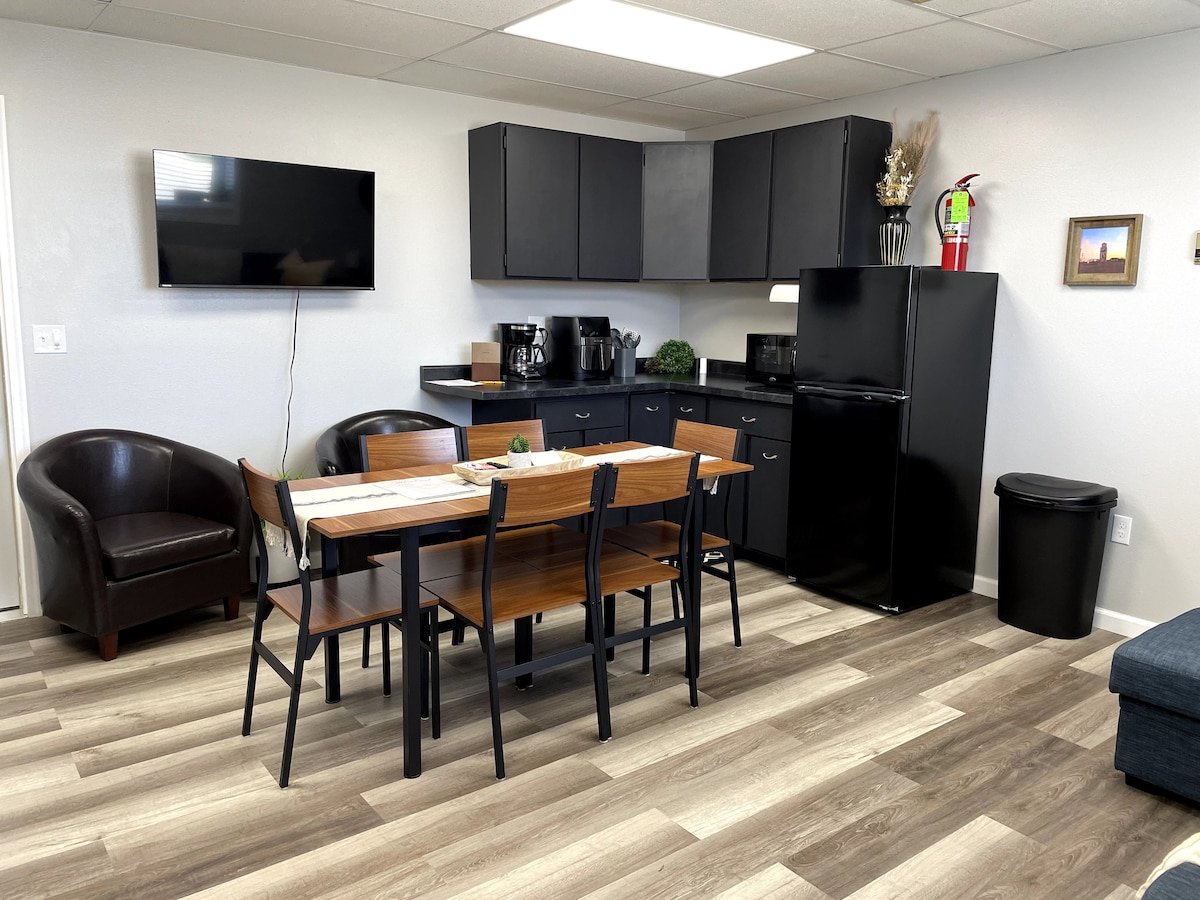 A modern kitchen area contains dark cabinets and a black refrigerator. A wooden dining table with metal chairs is set for six. A comfortable seating area includes two leather chairs. A flat-screen TV is mounted on the wall, enhancing the contemporary design.
