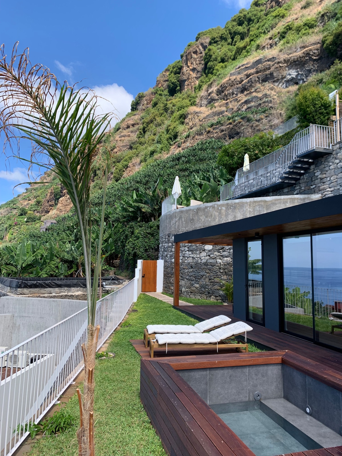 A serene outdoor space features a wooden deck surrounding a hot tub, overlooking mountainous terrain and the ocean. Two sun loungers are positioned nearby, providing a comfortable area for relaxation. Lush greenery and a few umbrellas can be seen in the background, enhancing the tranquil environment.