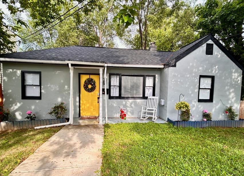 A charming house exterior features a bright yellow front door and a welcoming wreath. The façade includes white window trims and is complemented by flower boxes below the windows. Lush green grass surrounds a concrete pathway leading to the entrance, shaded by trees.