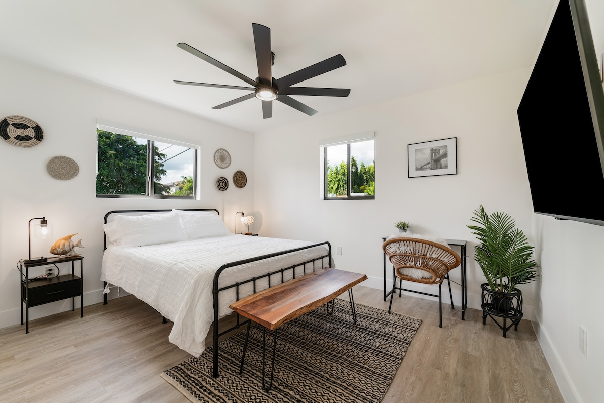 A well-lit bedroom features a spacious bed framed by two windows, allowing natural light to fill the space. A ceiling fan hangs above, while a wooden table and a chair provide a cozy sitting area. Decorative wall hangings add a touch of character.