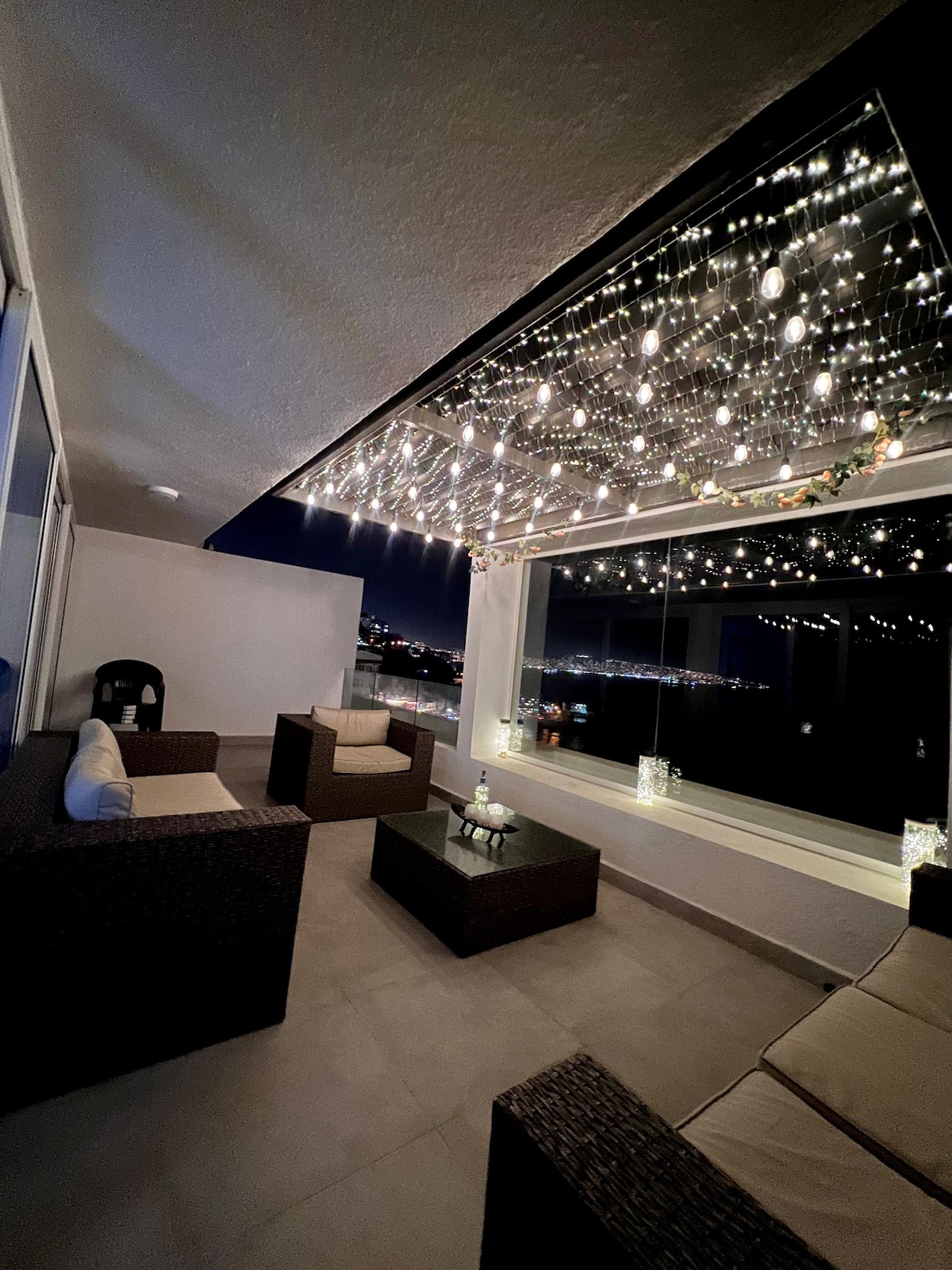 A modern terrace space features cozy seating arranged around a glass coffee table. Soft lighting from fairy lights hangs overhead, creating a serene ambiance. Large windows provide views of the surrounding area, enhancing the open and airy feel of the space.