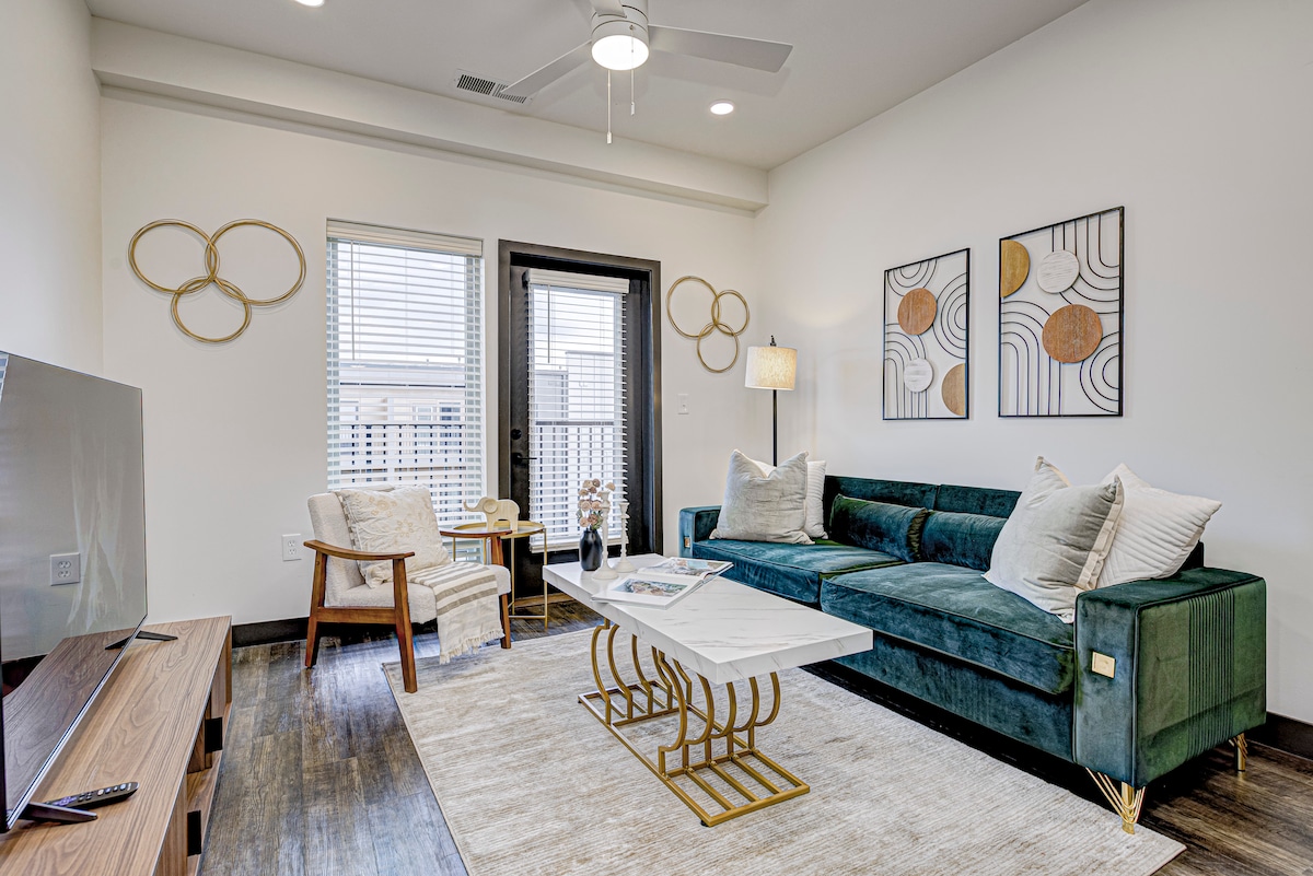A stylish living area features a plush green sofa paired with decorative pillows and a coffee table. Two modern wall art pieces enhance the decor, while large windows allow natural light to brighten the space. A comfortable chair adds an inviting touch.