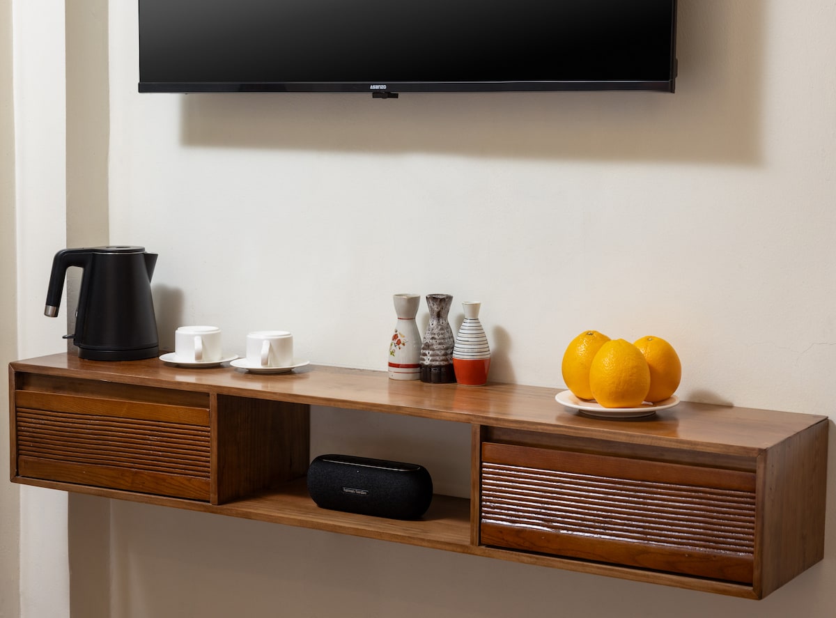 A wooden shelf is mounted to the wall, featuring a sleek electric kettle, two cups, and decorative ceramic pitchers. A bowl of oranges adds a pop of color. Below, a long, narrow speaker is positioned, completing the modern aesthetic of the room.