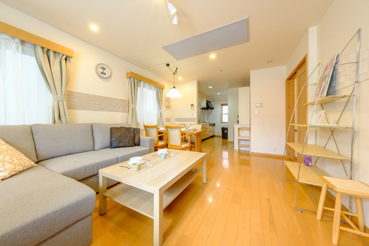A spacious living and dining area features a comfortable gray sofa complemented by wooden furniture. A rectangular table is set for casual dining with several chairs around it. Natural light flows through large windows, enhancing the warm wooden flooring.