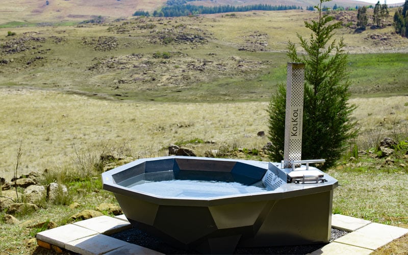 A modern wood-fired hot tub is positioned outdoors, surrounded by an expansive landscape. The tub's geometric design is highlighted against the natural backdrop of rolling hills and greenery, offering a serene setting for relaxation while enjoying the scenic views.